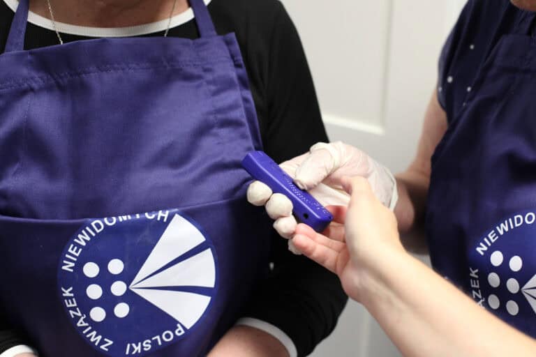 Dłonie (jedna w lateksowej rękawiczce) z niebieskim urządzeniem identyfikującym kolory. W tle widać fragment granatowego fartucha, na którym jest logo i biały napis: Polski Związek Niewidomych