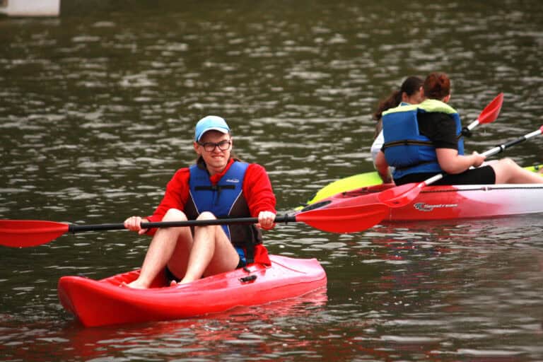 Na pierwszym planie, lekko z lewej w czerwonym kajaku płynie młody człowiek w okularach i błękitnej czapce typu „kaszkietówka”. Mężczyzna trzyma w dłoniach wiosło. Ma na sobie czerwoną bluzę i niebiesko-czarny kapok. Z prawej fragment innego biało-czerwonego kajaku