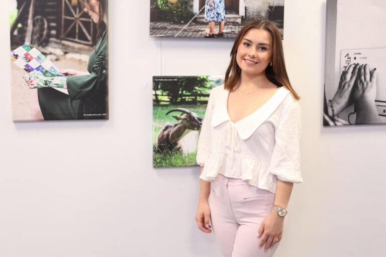 Paulina Muszyńska pozuje do pamiątkowej fotografii na tle zdjęć własnego autorstwa, wyeksponowanych na jasnej ścianie. Kobieta uśmiecha się do obiektywu