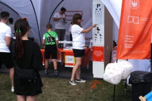 Od lewej kobieta ubrana na czarno, która bierze udział w badaniu wzroku. Z prawej kobieta wykonująca badanie wskazuje ręką na tablicę Snellena – do badania ostrości wzroku