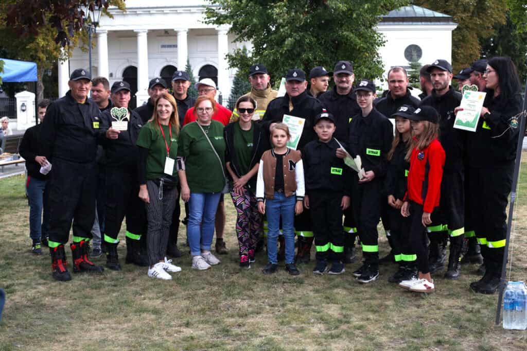 Grupowa fotografia pozowana. Ponad dwadzieścia osób, wśród nich strażacy i dzieci w kombinezonach strażackich. Niektórzy pozują z dyplomami i statuetkami. W tle jasny budynek z kolumnami