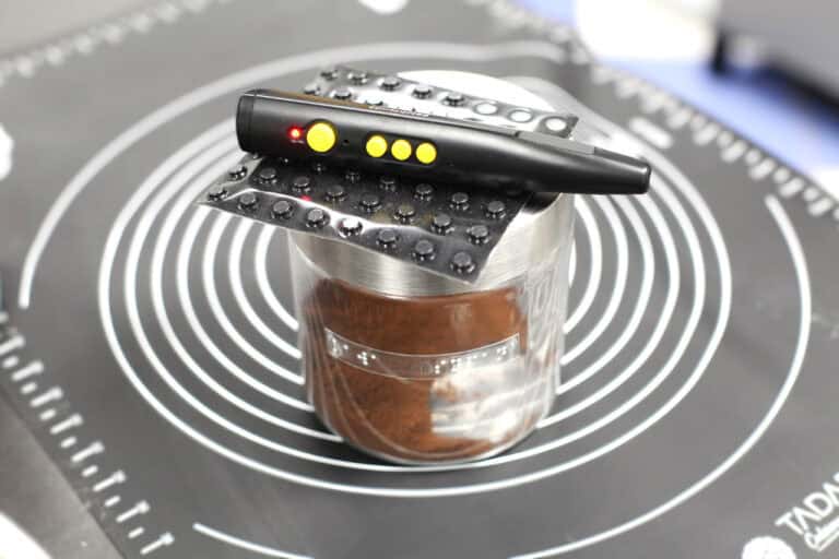 W centralnej części kadru, na czarnej macie do wyrabiania ciasta stoi słoik, z etykietą i nazwą produktu napisaną alfabetem Braille’a. Na nim leży naklejka Braille’a i i PenFriend – czarny z żółtymi przyciskami