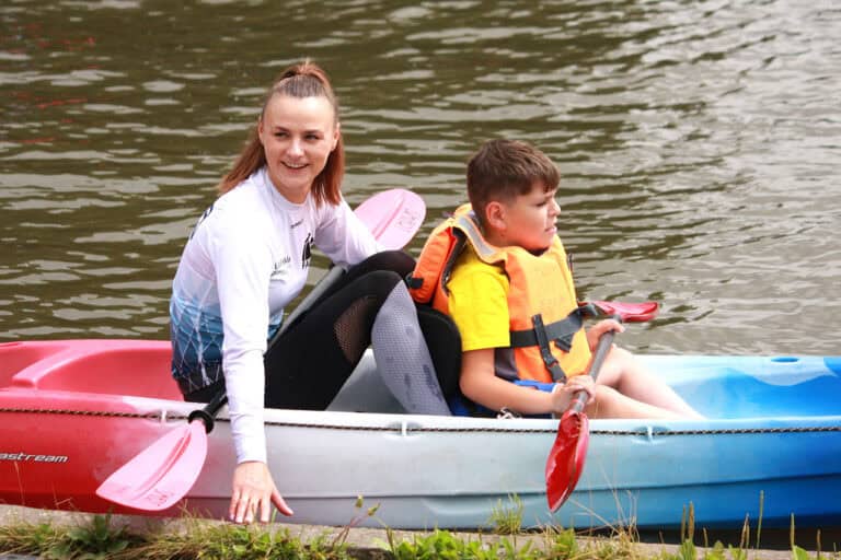 Wzdłuż brzegu czerwono biało niebieski kajak, a w nim dwie osoby: patrzący przed siebie chłopiec w pomarańczowym kapoku, za nim młoda kobieta, patrząca w bok i uśmiechająca się do kogoś poza kadrem. W tle woda