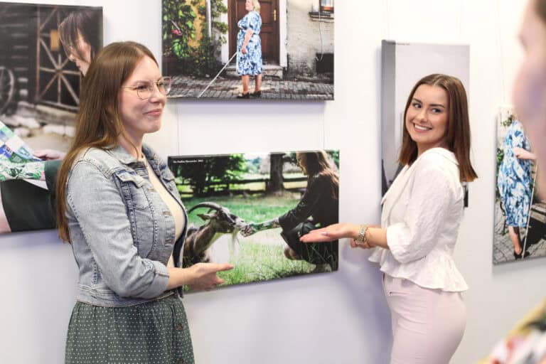 Dwie kobiety pozują na tle ściany z fotografiami, gestem dłoni wskazując na jedną z fotografii. Kobiety uśmiechają się