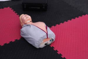 W centrum kadru na malinowo czarnej macie leży fantom – manekin do nauki pierwszej pomocy