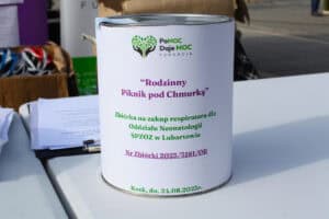 W centralnej części kadru biała puszka na datki. Puszka ma fioletowy napis: „Rodzinny Piknik Pod Chmurką” oraz numer zbiórki i zielony napis „Zbiórka na zakup respiratora dla Oddziału Neonatologii SPZOZ w Lubartowie” oraz logo Fundacji Pomoc Daje Moc