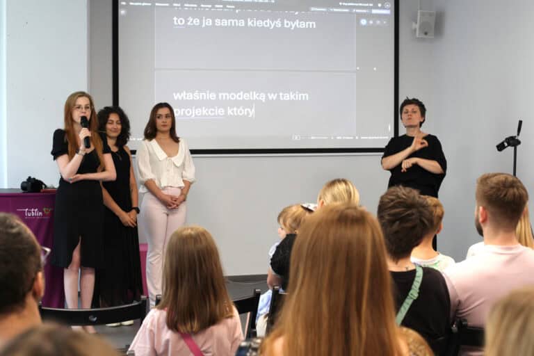 Wnętrze sali. Na pierwszym planie ludzie siedzący na krzesłach. Na drugim planie od lewej: trzy kobiety, z których pierwsza trzyma mikrofon. Z prawej tłumaczka języka migowego. W tle monitor z wyświetlanym na żywo tekstem