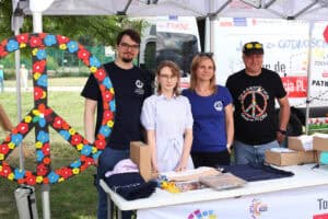 Od lewej duży znak pokoju – pacyfa, z wymalowanymi kolorowymi kwiatami. Do fotografii pozują cztery osoby prowadzące stoisko Tour de Konstytucja. Od lewej: mężczyzna, dwie kobiety, mężczyzna. Wszystkie osoby patrzą w obiektyw delikatnie się uśmiechając. W tle biały bus z napisem Jestem za godnością.