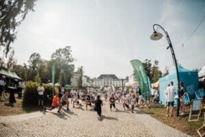 Duża grupa ludzi zgromadzonych przed jasnym budynkiem Zamku w Janowie Podlaskim. W centralnej części kadru uczestnicy biorą udział w grupowej rozgrzewce – wykonują wykroki. Rozgrzewka prowadzona jest przez instruktorkę stojącą przodem do nich. Większość osób ma na sobie sportowe stroje i numery startowe. Po bokach stoją namioty organizatorów oraz stoiska sponsorów wydarzenia. W tle znajdują się drzewa, lampy uliczne oraz słoneczne niebo, które dodaje wydarzeniu letniej atmosfery.