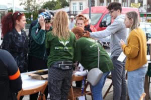 Punkt zapisu uczestników. Za stołem cztery osoby, dwie z nich ma na sobie zielone koszulki Fundacji Pomoc Daje Moc. Przed nim trzy kobiety, z których jedna mierzy niebiesko-biały kask rowerowy