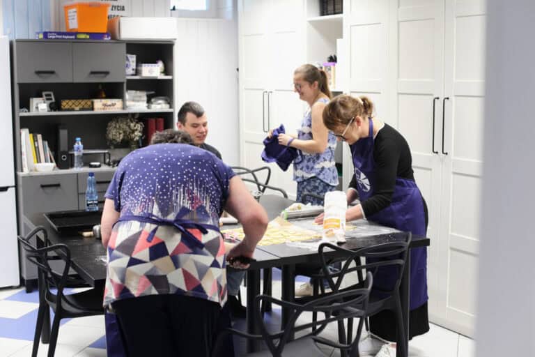 Cztery osoby (trzy stojące kobiety i siedzący mężczyzna) zgromadzeni wokół stołu, na którym znajdują się akcesoria do wypieków. Wokół nich białe, wysokie szafy kuchenne, a z lewej szary regał biurowy z szafkami zamykanymi i otwartymi półkami