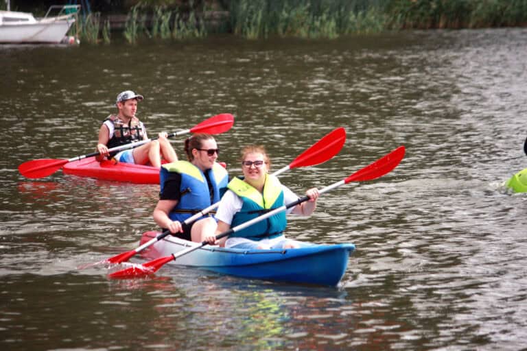 Na pierwszym planie dwie młode kobiety w kajaku. Obydwie mają na sobie niebiesko-żółte kapoki, trzymają wiosła. Za nimi, lekko z lewej w czerwonym kajaku płynie młody mężczyzna