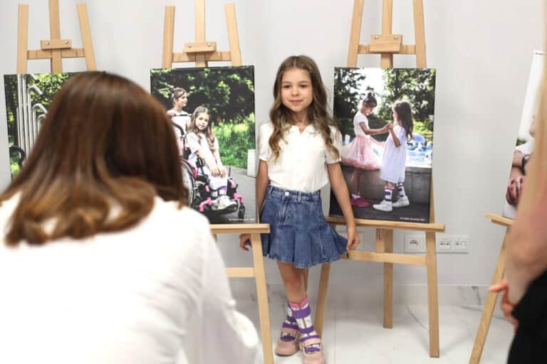 W centrum kadru dziewczynka pozuje między dwiema sztalugami, na których wyeksponowane są zdjęcia z jej podobizną. Dziewczynka pozując trzyma palcami rąbek dżinsowej spódniczki