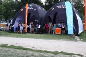 Na pierwszym planie pas drogi i trawy. Widok na dwa granatowe namioty KartoSfery. Przed namiotami i wewnątrz grupy osób. W tle zieleń drzew