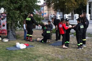 Kilkanaście osób, w tym dzieci i młodzież, większość w kombinezonach strażackich. Na trawie, na czarnych matach fantomy szkoleniowe do udzielania pierwszej pomocy. W tle zabudowania i zieleń drzew