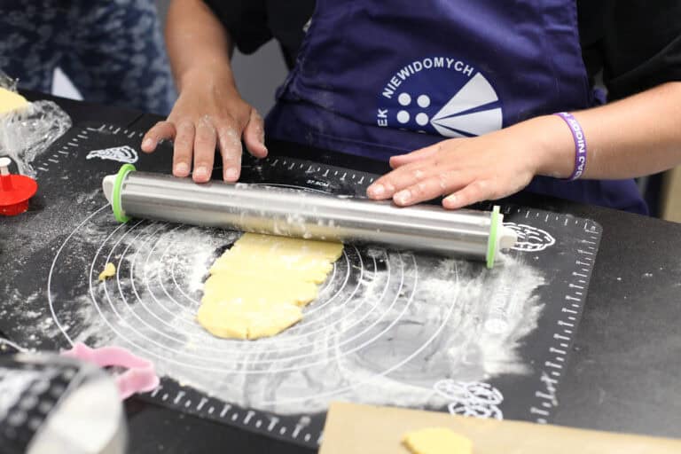 Zbliżenie na oprószoną mąką czarną matę do wyrabiania ciasta, na której leży podłużny wałkowany kawałek ciasta. Widać dłonie, które trzymają wałek, również oprószone mąką