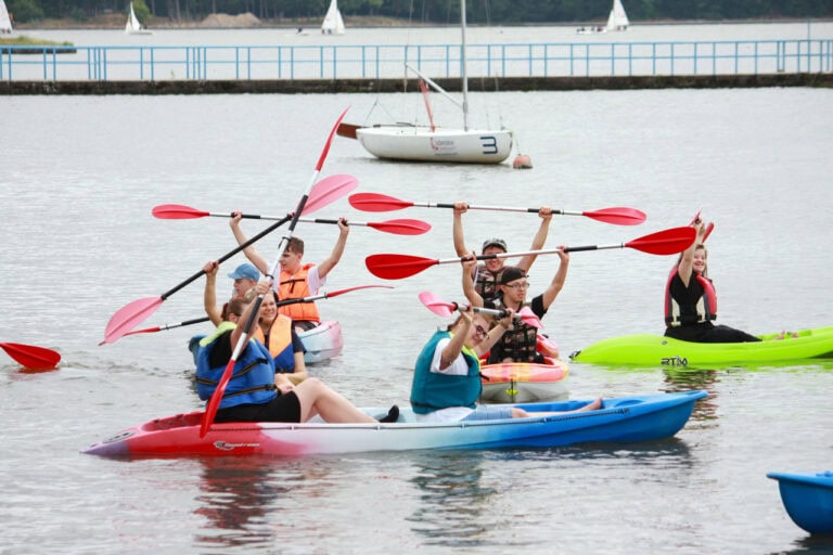 Cztery kajaki na wodzie. Osoby w nich siedzące unoszą ponad głowy wiosła. W tle łódź, za nią pomost. Na horyzoncie trzy żaglówki na tle lasu