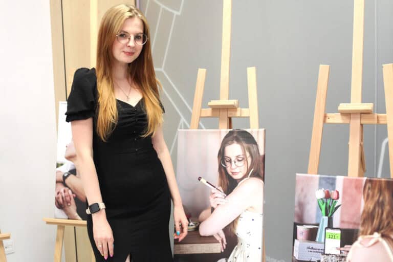 W centrum kadru przy sztaludze ze swoim zdjęciem pozuje młoda kobieta. Kobieta ma jasne proste włosy, okulary i jest ubrana w czarną sukienkę