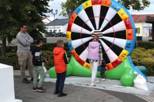 Na pierwszym planie mężczyzna z grupą dzieci. Stoją przed dużym kolorowym, dmuchanym kołem imitującym tarczę do rzutek. Koło ma pola czarno-białe i ponumerowane kolorowe pola. W tle zabudowania, po lewej stronie kadru fragment dużego drzewa