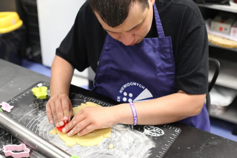 Mężczyzna pochylony nad rozwałkowanym kawałkiem ciasta, wykrawa za pomocą pomarańczowej foremki kształt ciastka. Ma na sobie czarną koszulkę z krótkim rękawem i granatowy fartuch Polskiego Związki Niewidomych, którego fragment logo jest widoczny na ujęciu