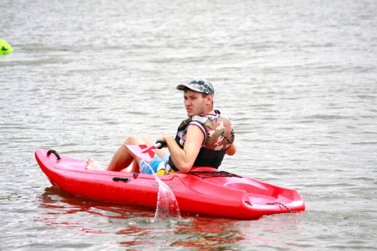 Młody mężczyzna płynie w czerwonym kajaku. Odwraca się przez ramię w kierunku fotografującego. Ma na sobie czapkę typu „kaszkietówka” i kapok z wzorem moro. Mężczyzna trzyma wiosło, z którego strumieniem spływa woda