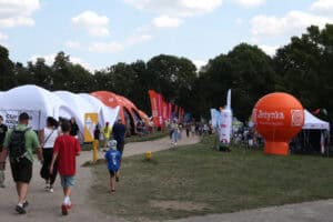 Widok ogólny na błonia, drogę między stoiskami i namiotami. Od lewej szereg namiotów, grupy osób, z prawej pomarańczowy balon reklamowy Jedynka Polskie Radio. W tle błękit nieba i zieleń drzew