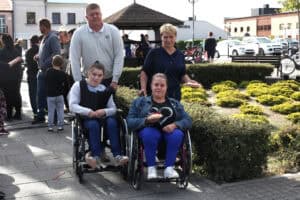 Pozowana fotografia rodzinna. Na pierwszym planie dwie młode kobiety siedzące na wózkach. Za nimi od lewej: mężczyzna i kobieta. Kobieta trzyma w lewej ręce gofra. W tle zgromadzeni ludzie i zabudowania