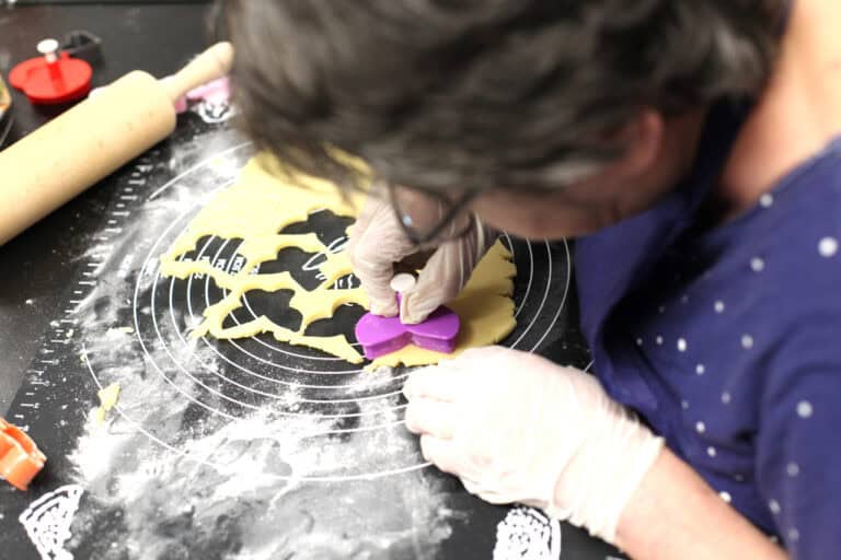 Rzut od góry na osobę pochyloną nad matą do wyrabiania ciasta, która w lateksowych rękawiczkach wyciska ciastka za pomocą fioletowej foremki w kształcie motyla. Mata jest w całości przysypana mąką, w lewym górnym rogu fragment drewnianego wałka do ciasta