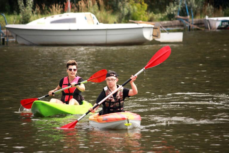 Dwa kajaki na wodzie. Od lewej płynie młoda kobieta w okularach przeciwsłonecznych, z jej lewej strony, wysunięty lekko do przodu kajak z młodym mężczyzną. Oboje trzymają wiosła. W tle: zacumowana przy brzegu łódź, za nią zieleń krzewów