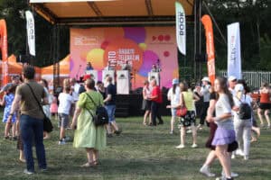 Na planie pierwszym około 30 osób tańczących przed sceną Lata z Radiem i Telewizją Polską. Na scenie czterech muzyków grających na instrumentach