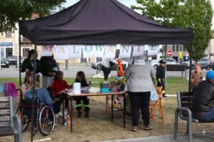 Kadr wypełnia stoisko "Malowanie twarzy". Stoisko ma czarny materiałowy dach w kształcie piramidy. Pod nim, na stoliku znajdują się akcesoria do malowania. Od lewej, obok stolika kobieta siedząca na wózku i kilka osób zgromadzonych wokół. W tle zabudowania. Po prawej drzewa