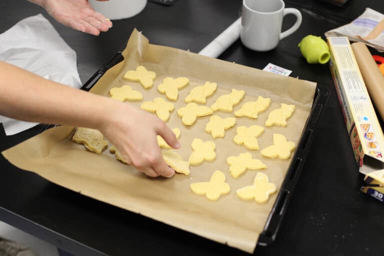 Zbliżenie na blachę piekarnika wyłożoną brązowym papierem do pieczenia, na którym znajdują się surowe ciastka w kształcie motyli i kotów. Od lewej dłoń, która kładzie ciastko na blachę