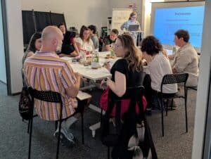 Uczestnicy warsztatów siedzą przy stole i pracują w parach, rozmawiając. W tle wyświetlana prezentacja i flipchart z napisem „Tu Otwieracz. Zapraszamy!”