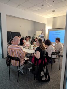 Trwa pierwszy blok warsztatowy, uczestnicy siedzą przy stole i pracują w parach, rozmawiając. W tle wyświetlana prezentacja i flipchart z napisem „Tu Otwieracz. Zapraszamy!”