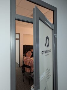 Uchylone drzwi wejściowe z logo Stowarzyszenia „Otwieracz”. W tle kilku uczestników spotkania.