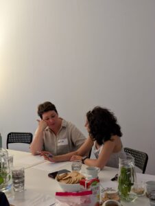 Przy stole siedzą dwie kobiety – po lewej reprezentantka Teatru Osterwy, a po prawej prezeska Fundacji Głuche Miasto, wspólnie pracują podczas jednego z zadań. Kobieta po lewej słucha z uwagą. Na stole ustawiono naczynia i przekąski.