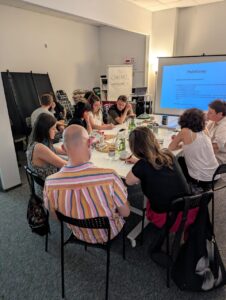 Widok na salę warsztatową w przestrzeni Stowarzyszenia „Otwieracz” i uczestników spotkania siedzących w okręgu przy stole.