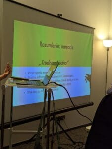 Kadr skierowany na laptopa i znajdujący się w tle ekran multimedialny z wyświetlonym slajdem prezentacji. Na nim napis: „Zrozumieć emocje. Trudno = nudno”.