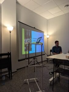 Laptop stojący na stoliku z regulowaną wysokością. W tle ekran multimedialny z wyświetloną prezentacją. Po prawej stronie przy ścianie widoczny tłumacz PJM, Łukasz Grzesiuk.