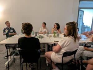 Uczestnicy spotkania siedzą przy stole. Część z nich patrzy w kierunku znajdującej się poza kadrem osoby prowadzącej, a część w kierunku tłumacza języka migowego (z lewej strony kadru).