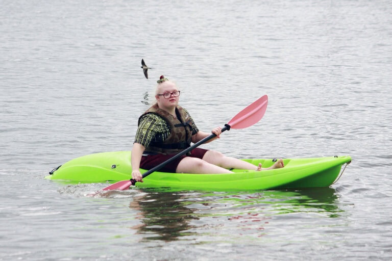 W centralnym punkcie kadru w jaskrawo zielonym kajaku płynie młoda kobieta w okularach. Kobieta zanurza w wodzie wiosło. Nad jej głową, z lewej strony ptak uchwycony w locie