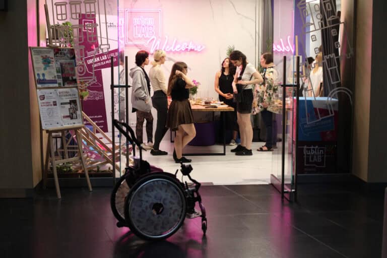 Kadr zrobiony poza wnętrzem galerii. Na pierwszym planie, w półmroku, przed wejściem do galerii wózek. W tle jasne wnętrze galerii, kilka osób pogrążonych w rozmowie. Z lewej, na ścianie różowy neon Urban Lab Welcome