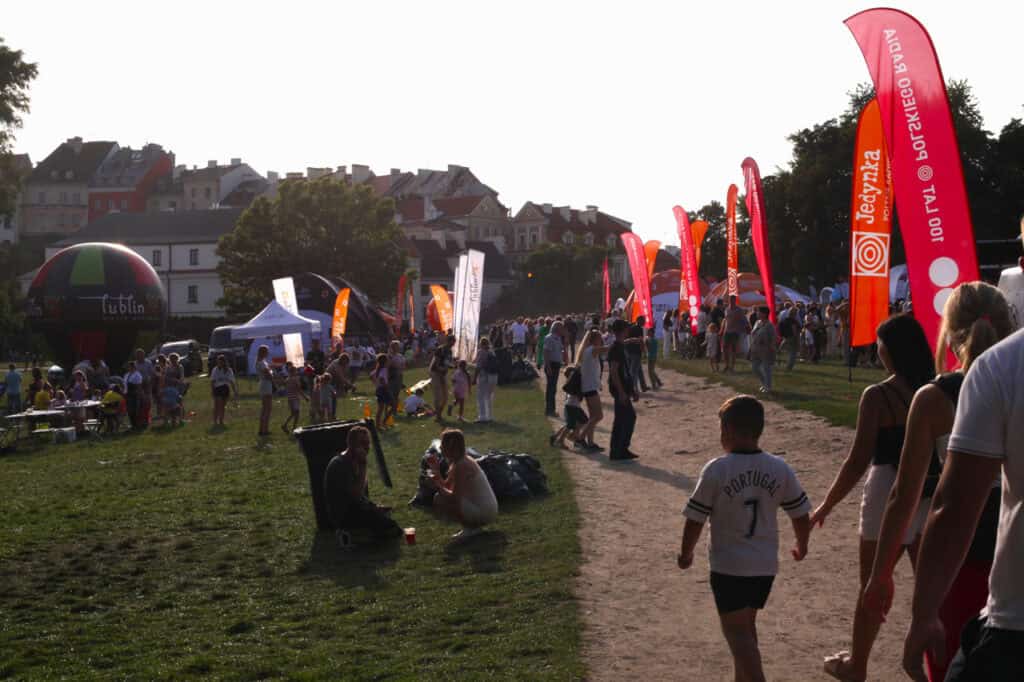 Widok na błonia w kierunku Starego Miasta. Na pierwszym planie grupy ludzi idących drogą, odpoczywających na błoniach. Z prawej wysokie pomarańczowe chorągwie Jedynka Polskie Radio. W tle dachy i kamienice Podwala oraz Starego Miasta