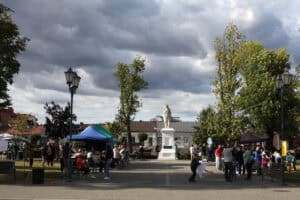 W centralnej części kadru pomnik św. Heleny. Po jego obu stronach rzędy stoisk z kolorowymi parasolami, przy których zgromadzone są grupy osób. Po lewej latarnia. W tle wysokie drzewa i niebo niebiesko-szare wypełnione chmurami