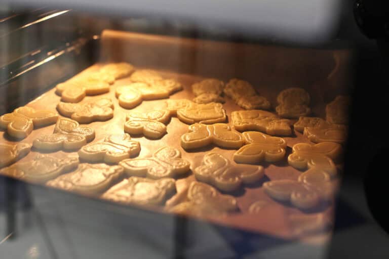 Wnętrze piekarnika, ujęcie przez szybę. Na blasze ciastka w trakcie pieczenia. Mają kształt motyli i kotów, delikatnie połyskują w świetle piekarnika