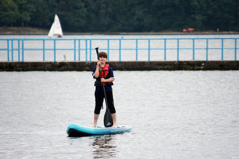 Na pierwszym planie na niebieskiej desce typu Sup stoi młody człowiek, trzyma w rękach wiosło. Za nim pomost z niebieskimi barierkami. W tle, na wodzie z lewej żaglówka. Na horyzoncie linia lasu