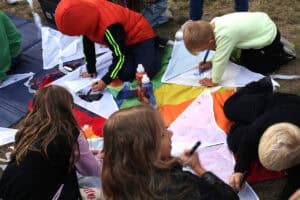 Rzut z góry. Na kolorowej macie siedzi pięcioro dzieci pochylonych nad białymi latawcami. Dzieci są w trakcie zdobienia latawców kolorowymi markerami