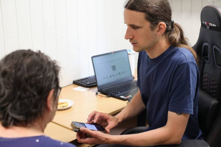 Zajęcia z tyfloinformatyki. Mężczyzna siedzący przy biurku (rzut z boku), trzyma w rękach telefon komórkowy. Postawa ciała zwrócona do osoby, która znajduje się w większości poza kadrem