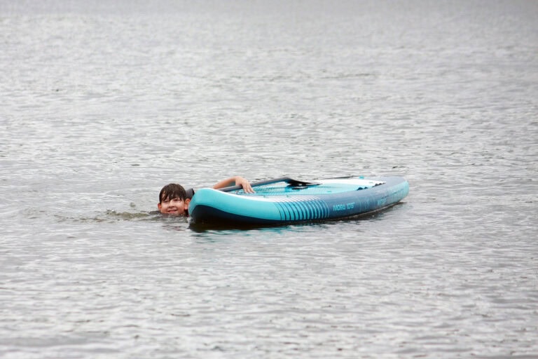 W centrum kadru, na wodzie niebieska deska typu Sup. Opiera się o nią ręką młodzieniec, któremu z wody wystaje tylko głowa. Chłopiec uśmiecha się