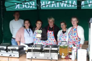 Pozowana fotografia pamiątkowa. We wnętrzu stoiska Koła Gospodyń Wiejskich pozuje sześć osób. Od lewej mężczyzna, kobieta, kobieta w okularach i kolorowym, wzorzystym fartuchu trzyma w rękach statuetkę. Kolejno trzy kobiety w kolorowych fartuchach. Przed nimi dwie duże gofrownice i duży słój z pomarańczową lemoniadą